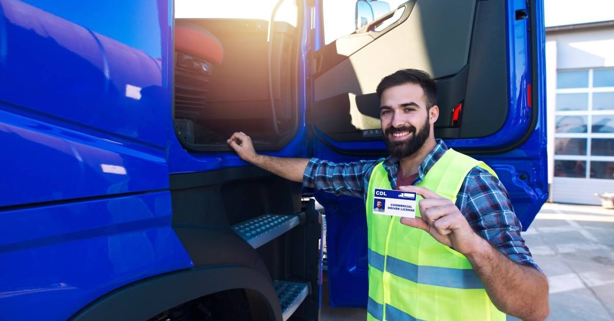 A bearded man wearing a high-visibility vest stands in the open doorway of a blue truck while smiling and holding up a CDL.