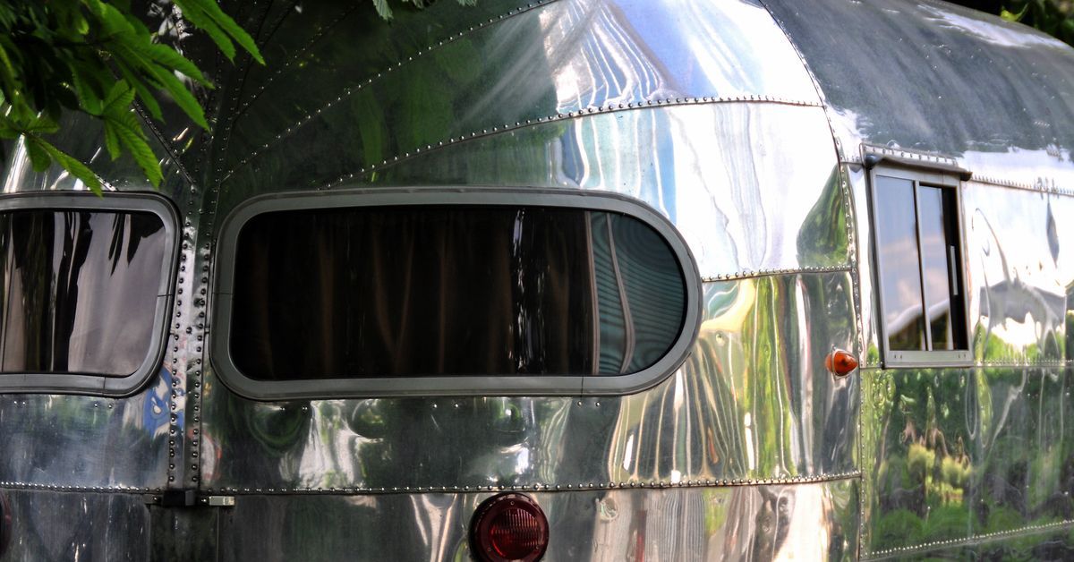 An airstream trailer parked outside next to a tree. The trailer is made of metal and has two red lights.
