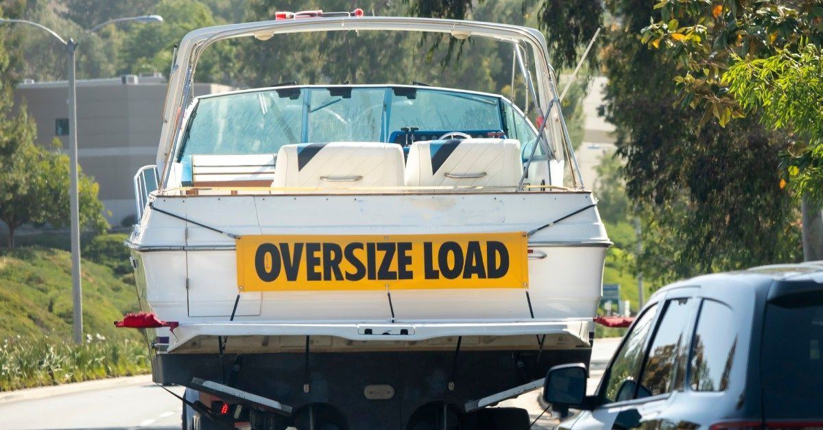 A white boat with a sign that says “oversize load” is transported on a roadway in front of a black car.
