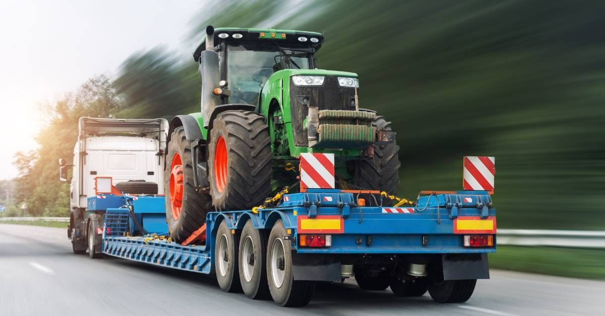 A white semi-truck with a blue trailer carrying a green tractor. The truck is racing down the highway, blurring its surroundings.