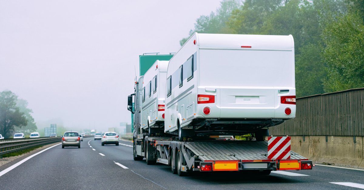 Two white camper trailers being transported by a semi-truck. The truck is moving down a highway behind other cars.