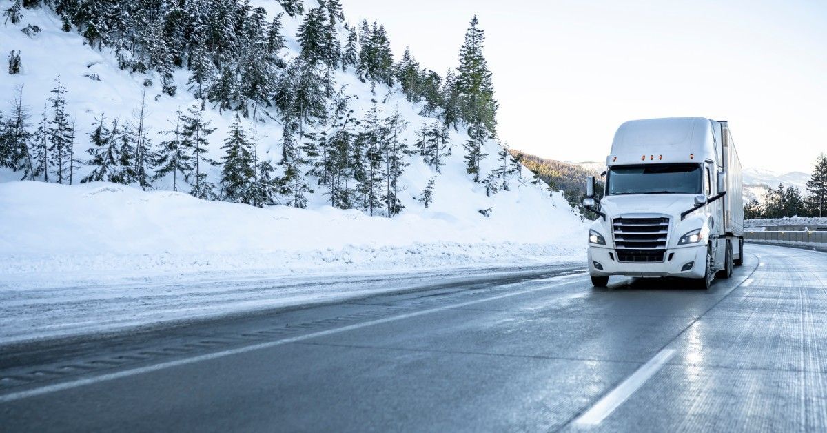 A white semitruck drives on a highway. The hill to the side of the highway is covered in trees and snow.