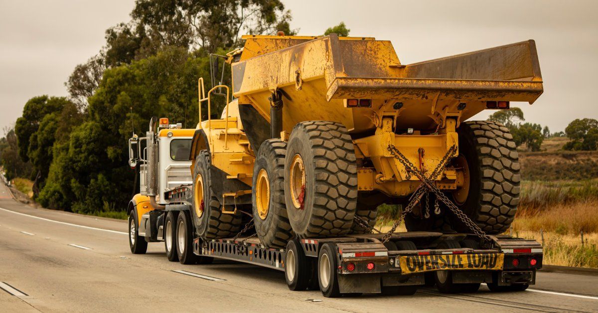 A dirty, yellow dump truck on the highway. The back of the trailer has a sign reading “oversize load.”