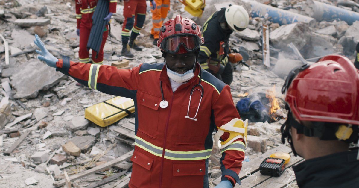 A man in a red uniform and helmet talking to another person while gesturing toward his right. Behind him is a mound of rubble.