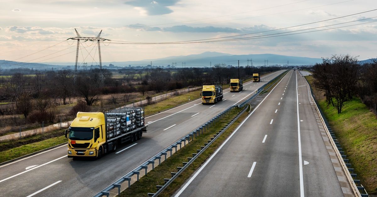 Four yellow trucks carry silver gas canisters on flatbeds while driving in one lane of a four-lane highway.