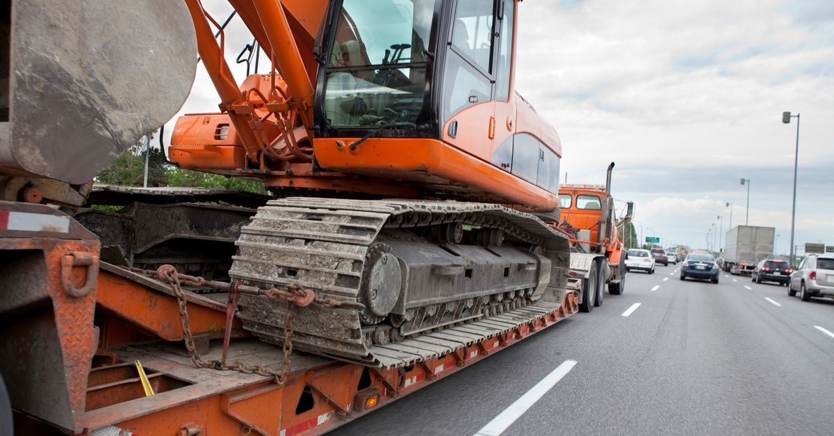 A side view of a semi-truck driving down a highway with an oversized construction machine. Other cars are ahead of the semi.