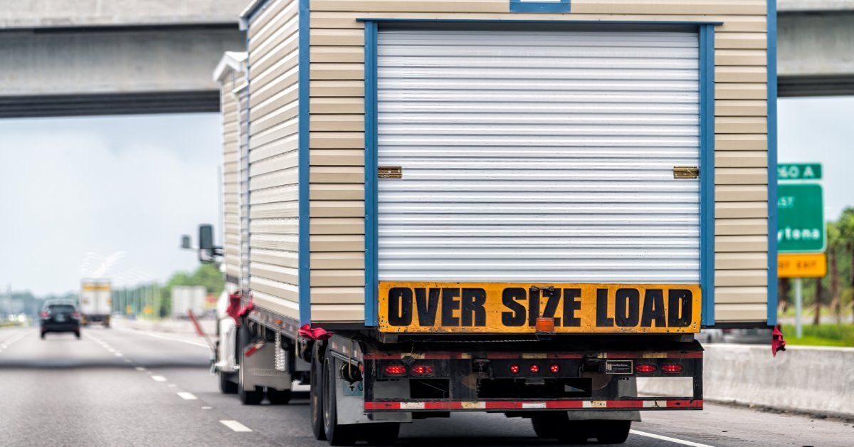 A hauler truck travels down the interstate with a tiny home. The back of the home has a banner that says
