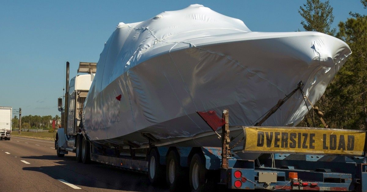 A large boat wrapped in white shrink wrap on the back of a trailer. The semi hauling the boat has a sign that says 