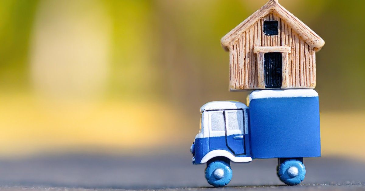 A blue toy truck with oversized wheels carries a small wooden house on its bed. The background is blurry.