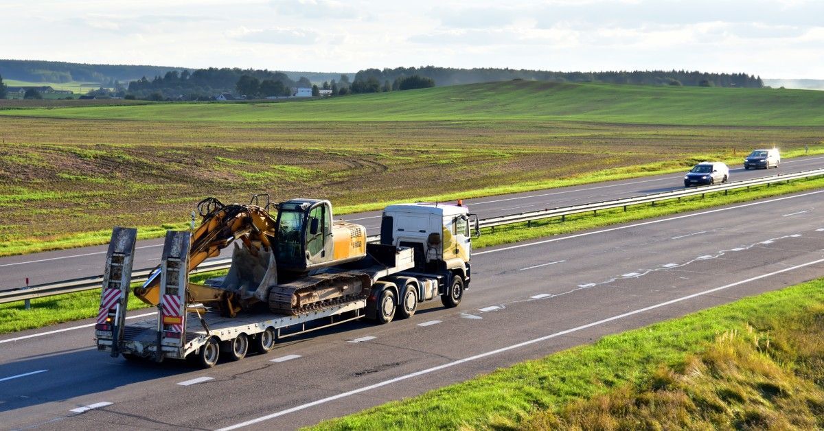A trailer truck with a long platform transports a large excavator across a highway. Other cars drive on the other side of the road.