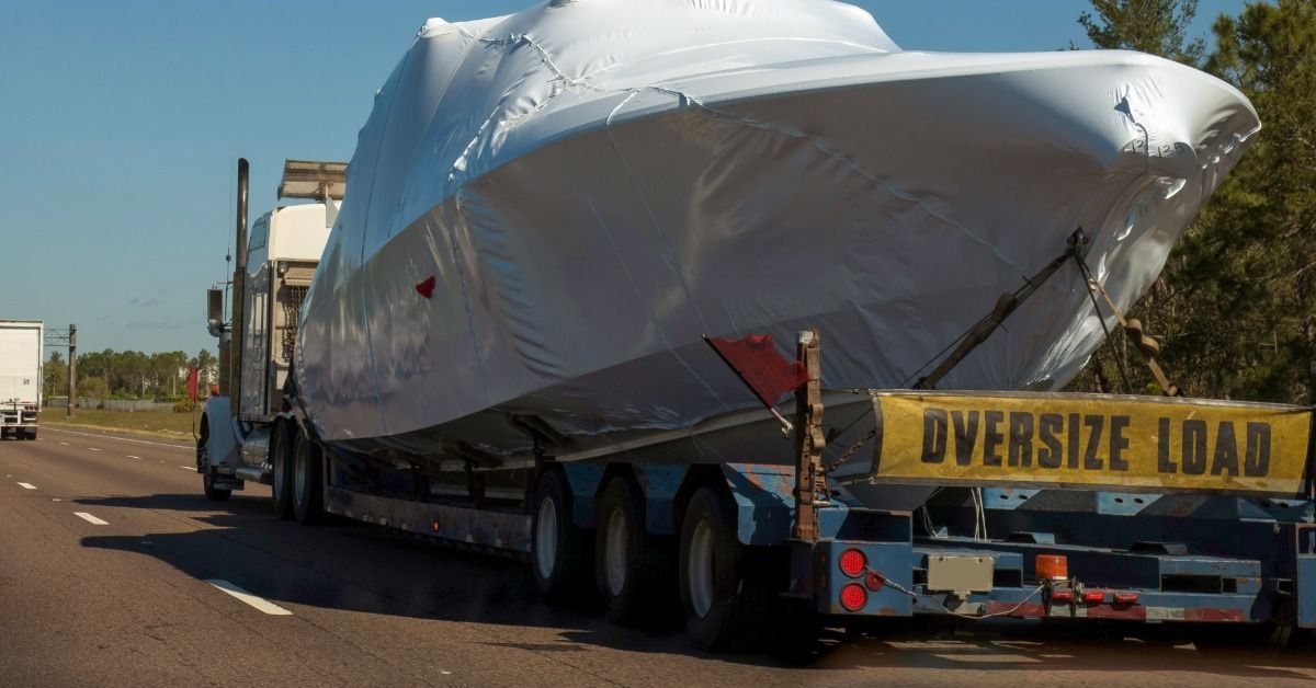 A yachy covered in white shrink wrap is strapped to the back of a semi-trailer with a banner saying