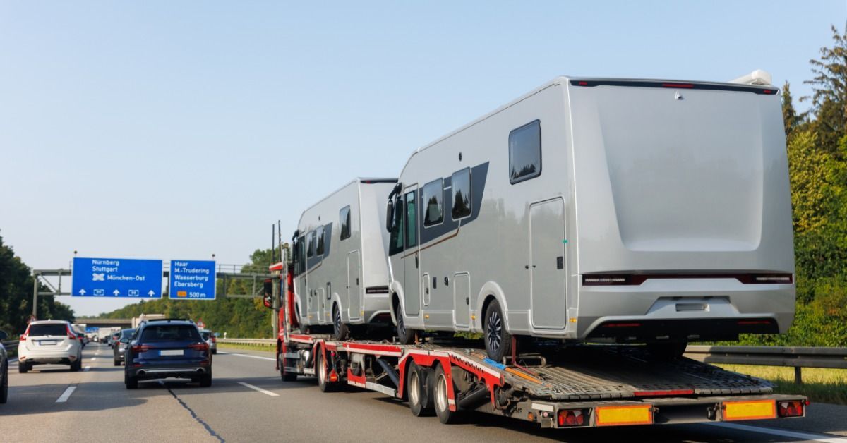 A semi with a long trailer transports two large grey RVs. Other cars drive along the highway too.