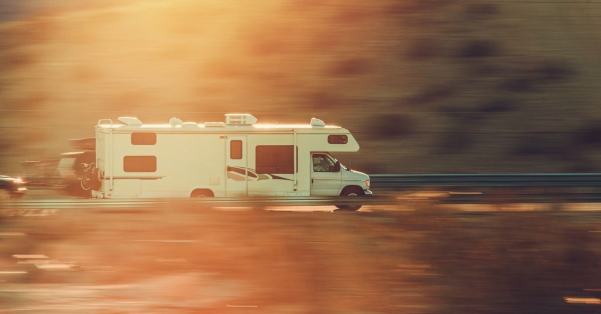 A white motorhome driving on a highway with blurred pavement, roadside grass, and green trees behind it.