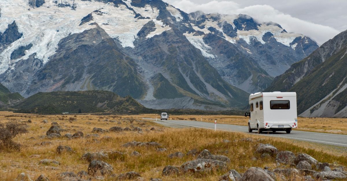 A white motorhome driving along a mountain road beside a river, with snow-covered peaks in the distance.