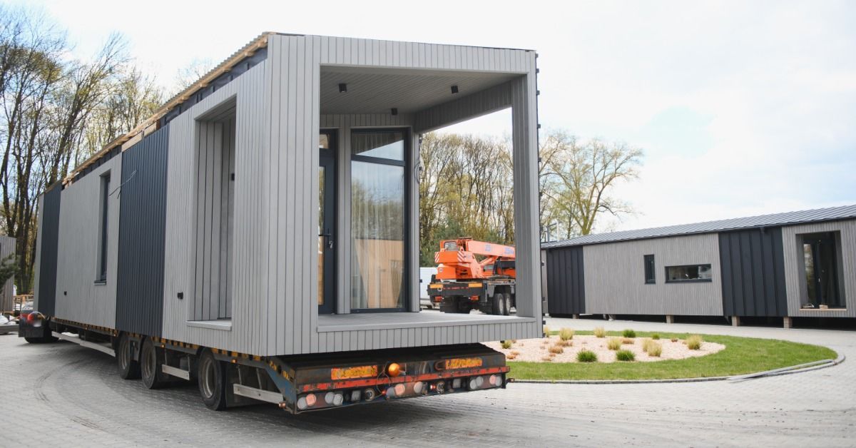 The back of a tiny home on a long flatbed trailer. The tiny home has tall glass windows and a small porch built into the back.
