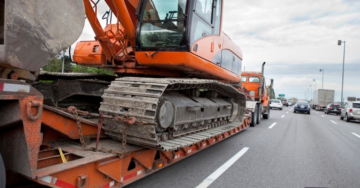A side view of heavy construction equipment on the back of a semi-trailer driving in three-lane highway traffic.