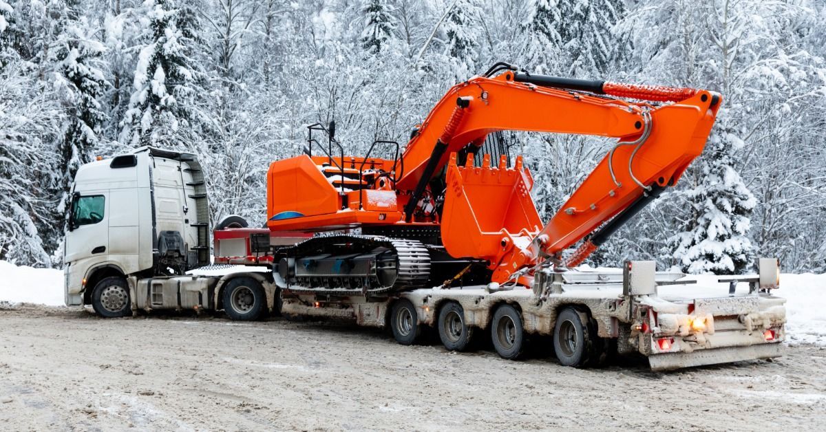 A large semi carries a bright orange excavator strapped to a trailer. The road and trees behind the semi are covered in snow.