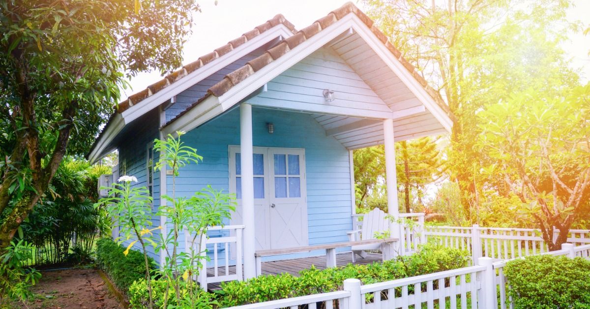 A tiny home with light blue siding and a white picket fence. There is a small porch with a double front door.