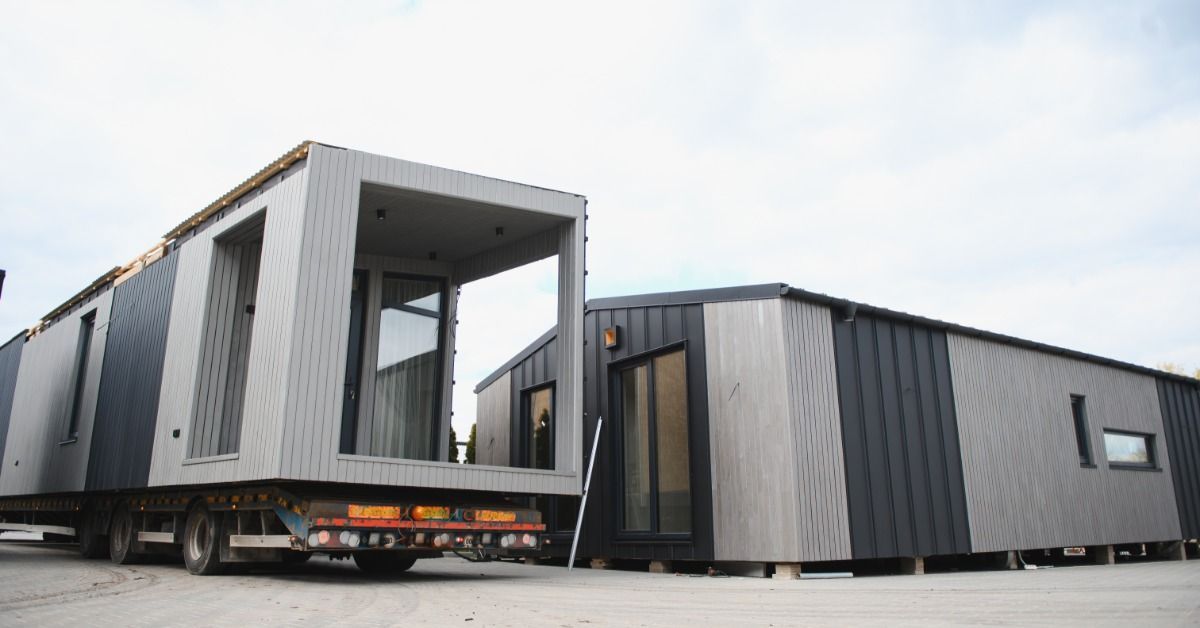 A modular tiny home with dark and light grey siding on the back of a flatbed trailer. Another home is next to it.