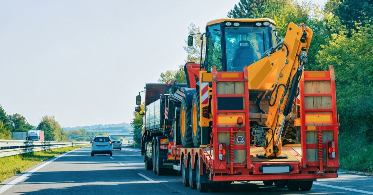 Tractor rides on a low trailer behind a semi on a highway. Greenery and distant hills frame the road