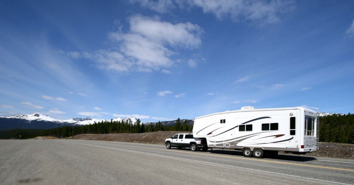 A pickup truck tows a white travel trailer along a highway. Forested mountains rise beneath a cloudy sky.