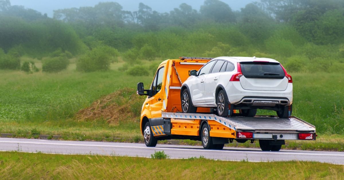 A flatbed tow truck carries a white SUV along a rural road through a green countryside transportation route.