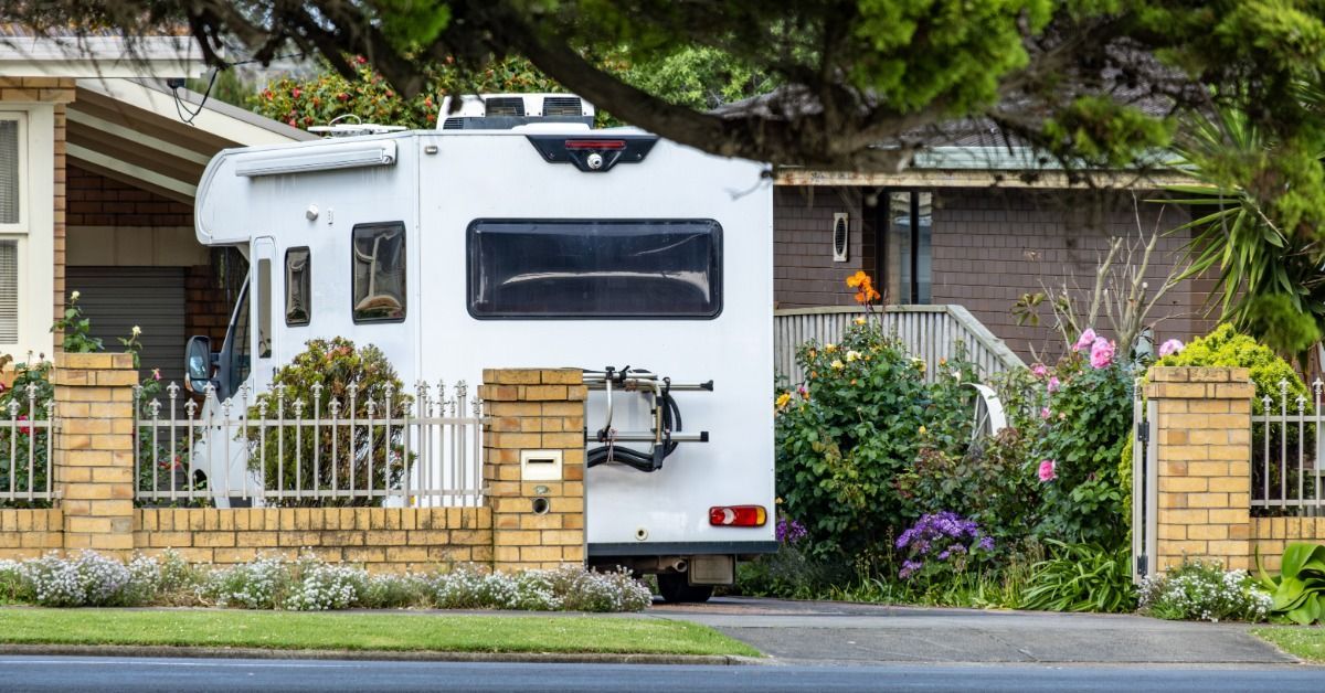 White motorhome parked in a residential driveway surrounded by a brick fence, landscaping, and overhanging trees.