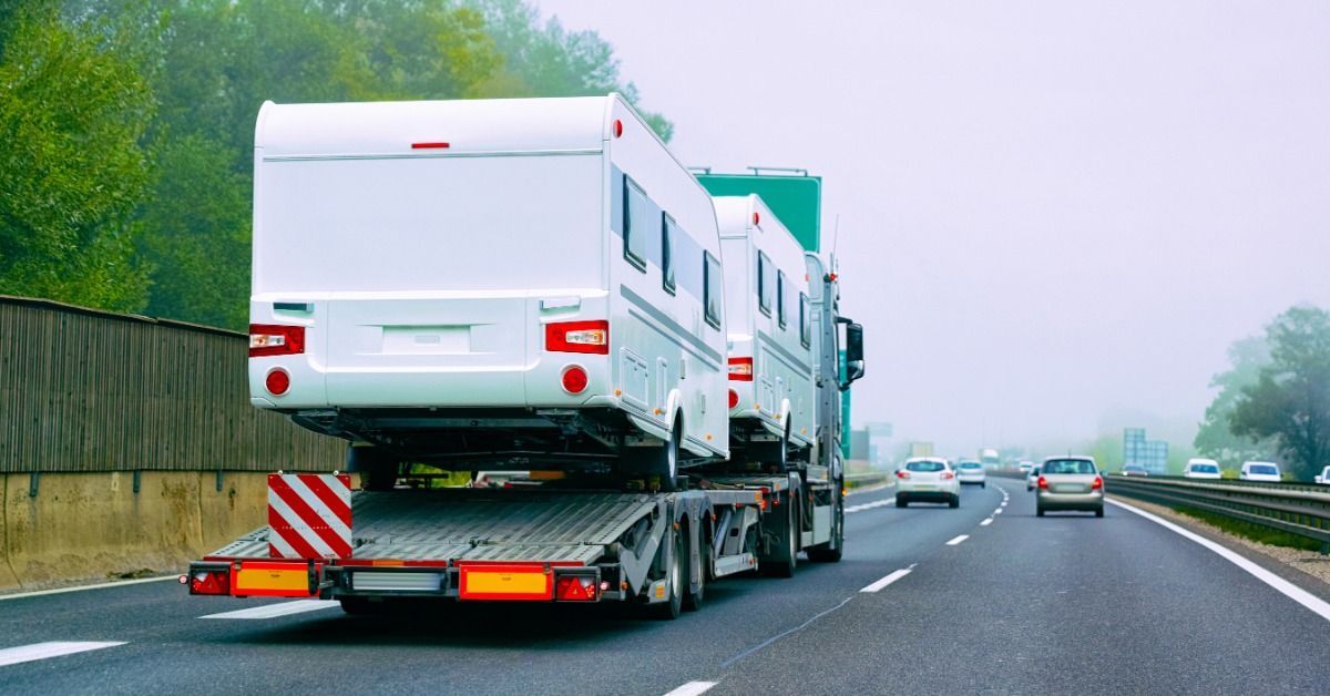 Two white motorhomes secured on a flatbed transport truck traveling along a highway with other vehic