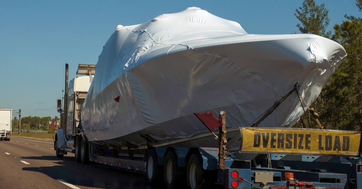 A truck carries a boat covered by a white tarp with an “OVERSIZE LOAD” banner on the back along a tree-lined road.