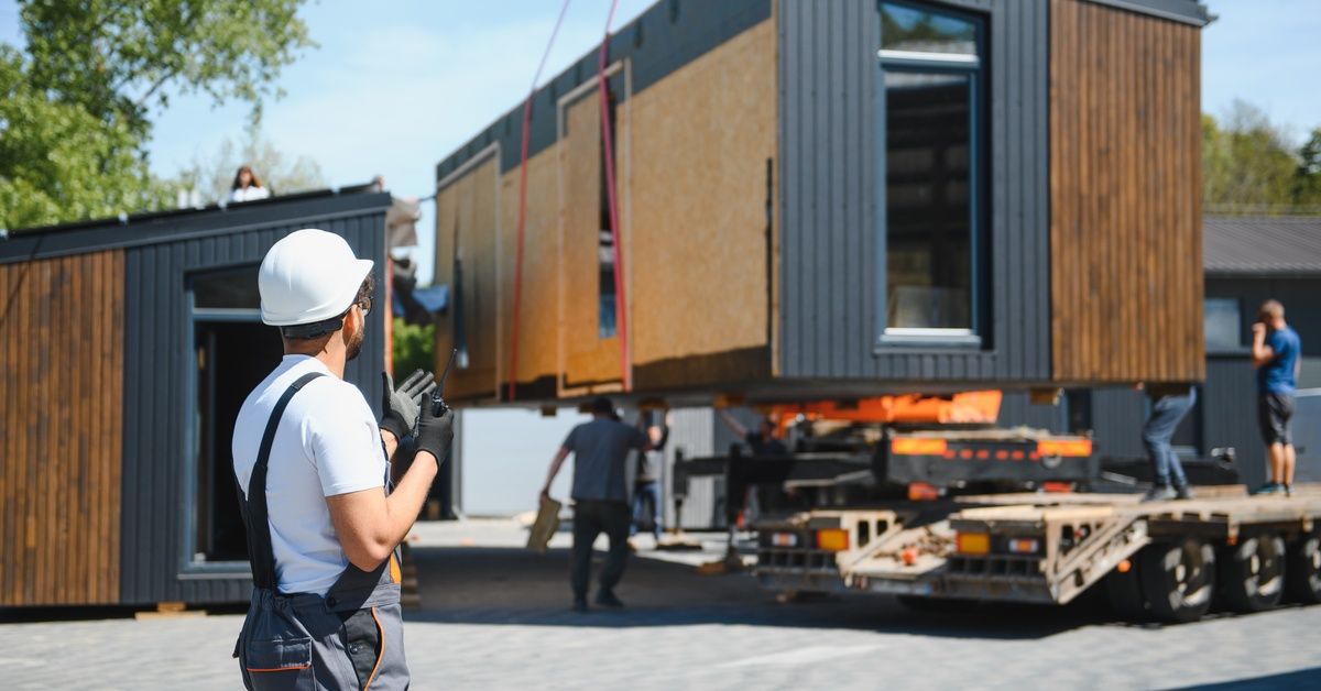 An orange crane lifts a tiny modular house onto a truck, as a man in gray overalls holds a walkie-talkie.