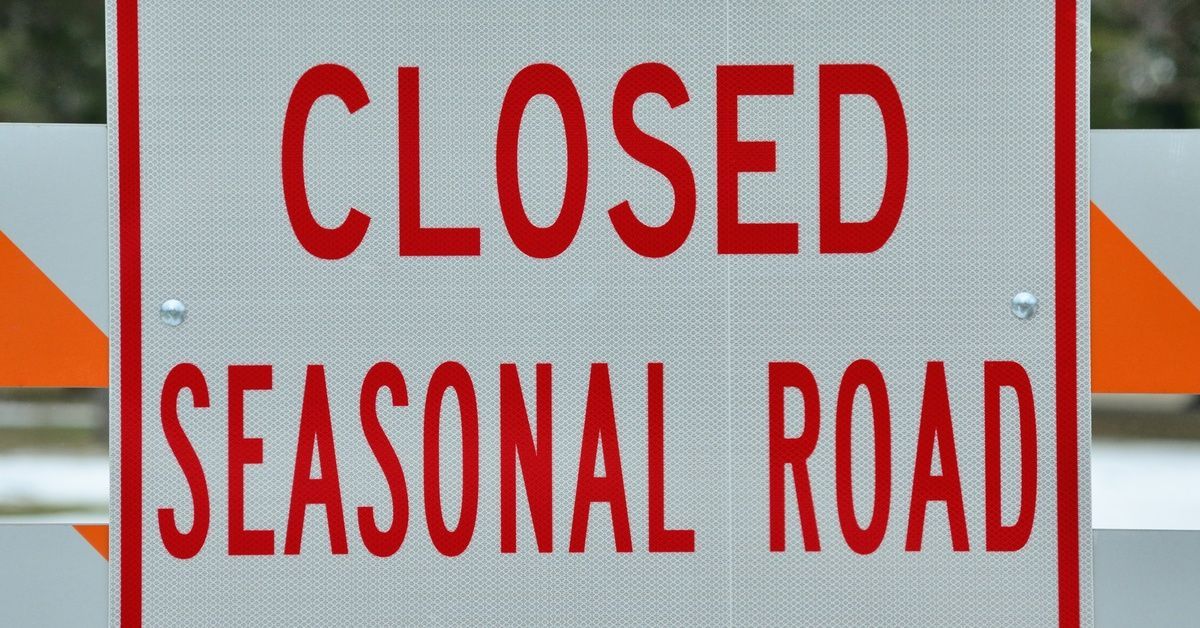 A white sign with reflective text reading “Closed” and “Seasonal Road” is mounted to three striped barrier posts.