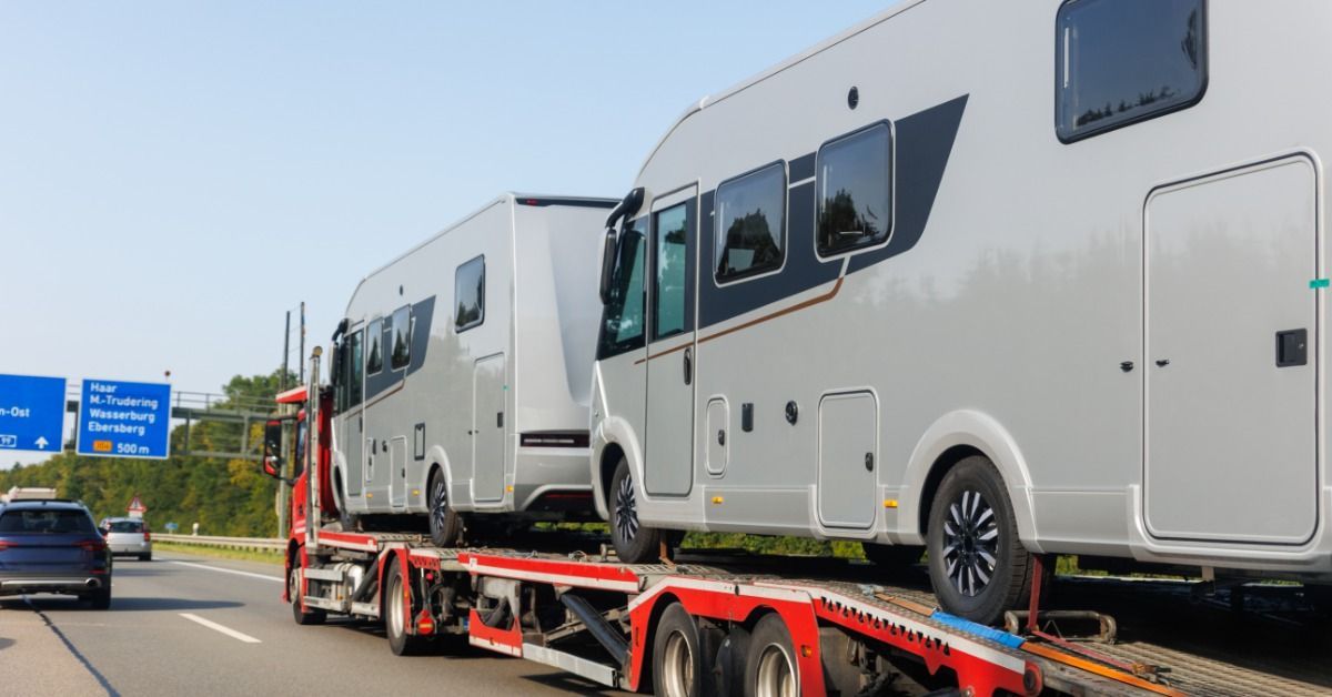 Two motorhomes are secured on a transport trailer while traveling along a highway.