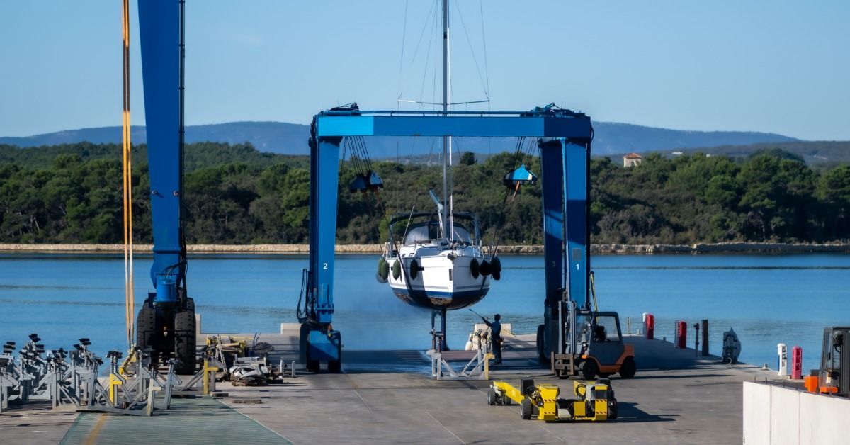 A marina travel lift raises a sailboat from the water as workers prepare the vessel for transport on land.