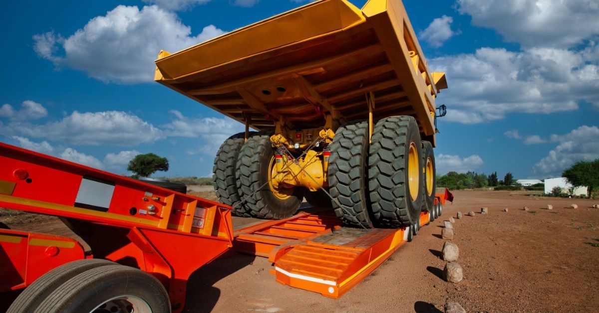 A large dump truck sits on a lowboy trailer positioned on an unpaved surface at a heavy equipment job site.