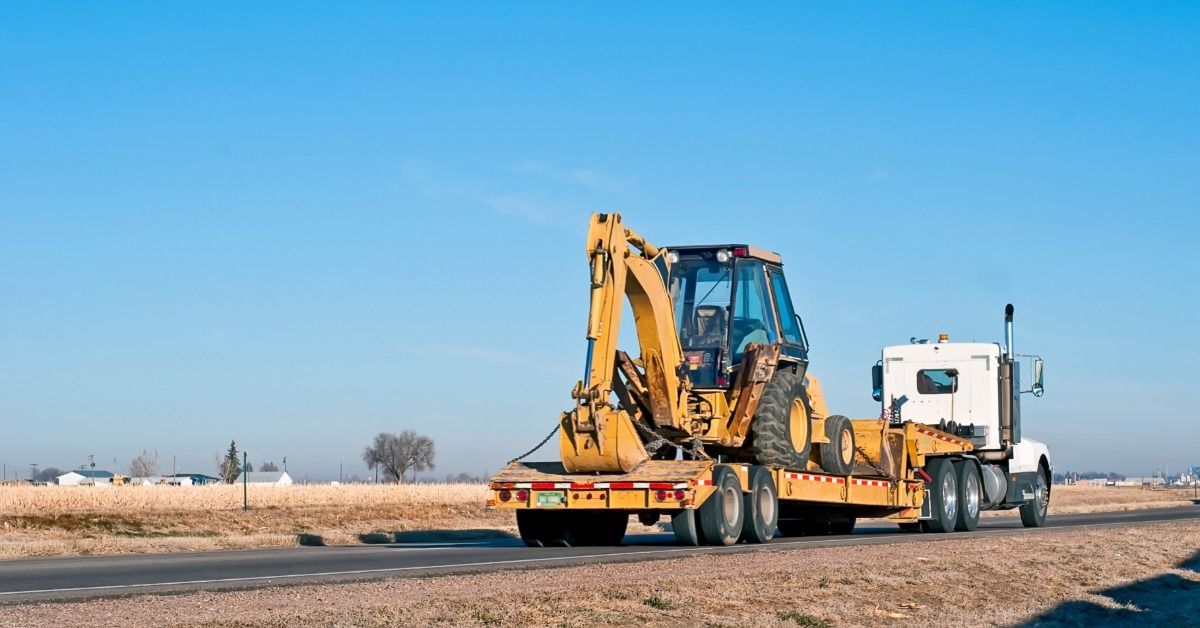 A backhoe loader is secured on a flatbed trailer as a transport truck carries it along a highway.