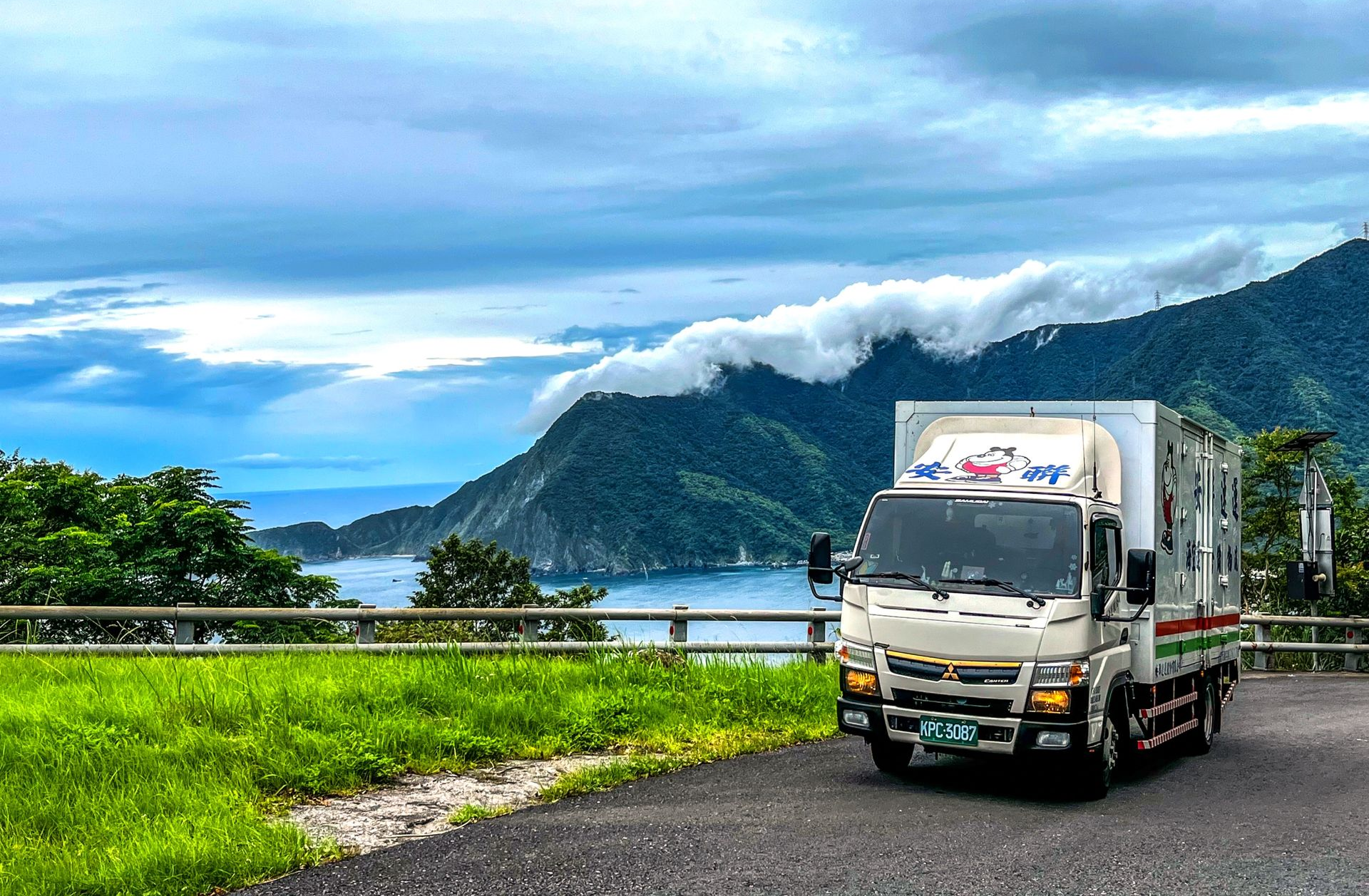 一輛白色卡車行駛在山路上，旁邊是湖泊，背景是綠色的山丘和陰雲密布的天空。