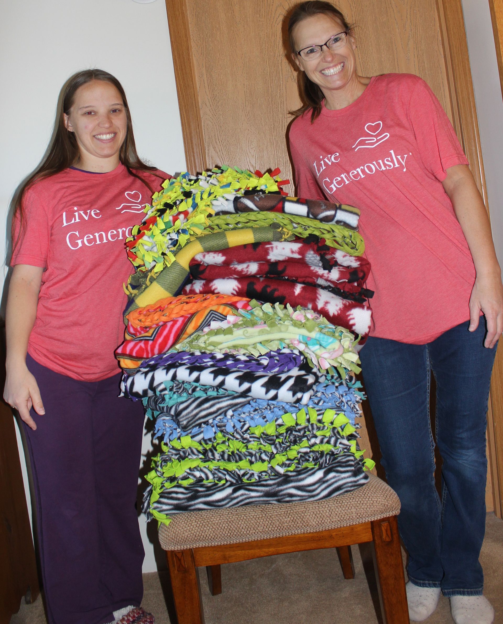 Two women in red shirts and jeans with a large stack of colorful knotted fleece blankets.