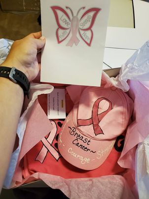 Person holding a gift box containing pink breast cancer awareness items: hat, shirt, and card with butterfly graphic.