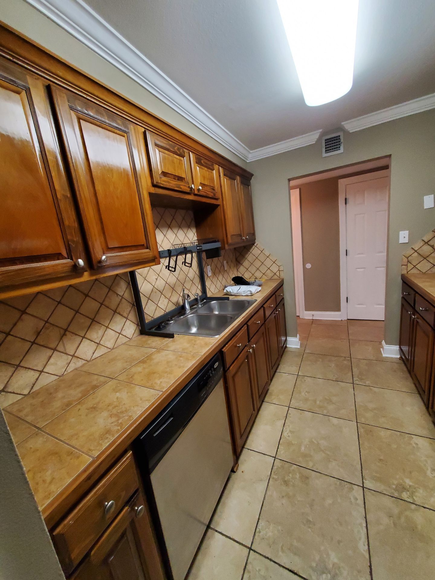 Narrow kitchen with wood cabinets, tan tile counters and floor, black sink, and a doorway at the far end