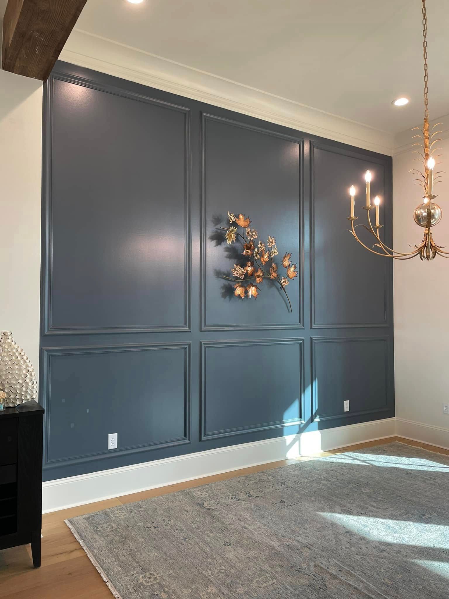 Dark blue paneled wall with floral wreath, chandelier, and gray rug in a bright room