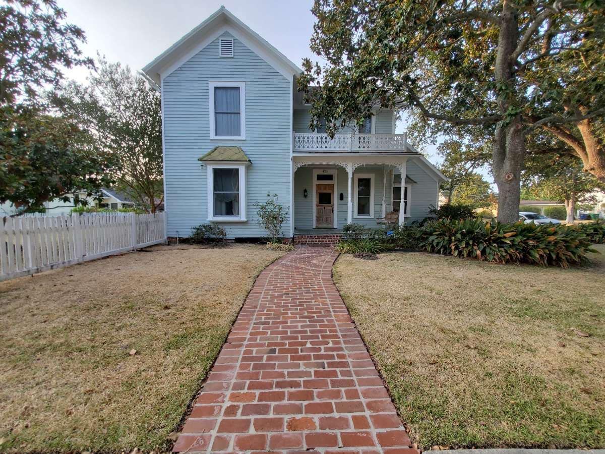 Blue house with a red brick walkway leading to the front door, bordered by a dry lawn and trees.