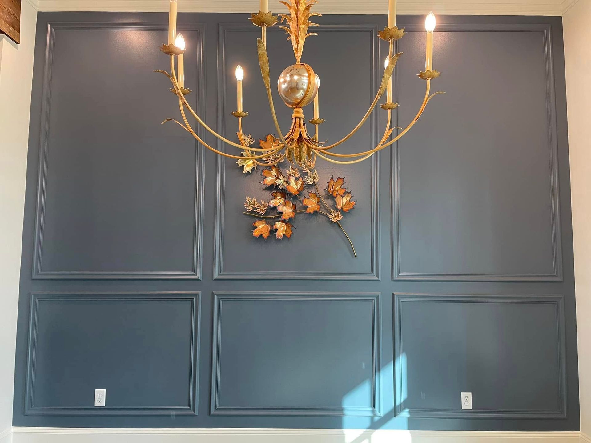 Blue paneled wall with gold chandelier and autumn floral decor, sunlight streaming in from the right.