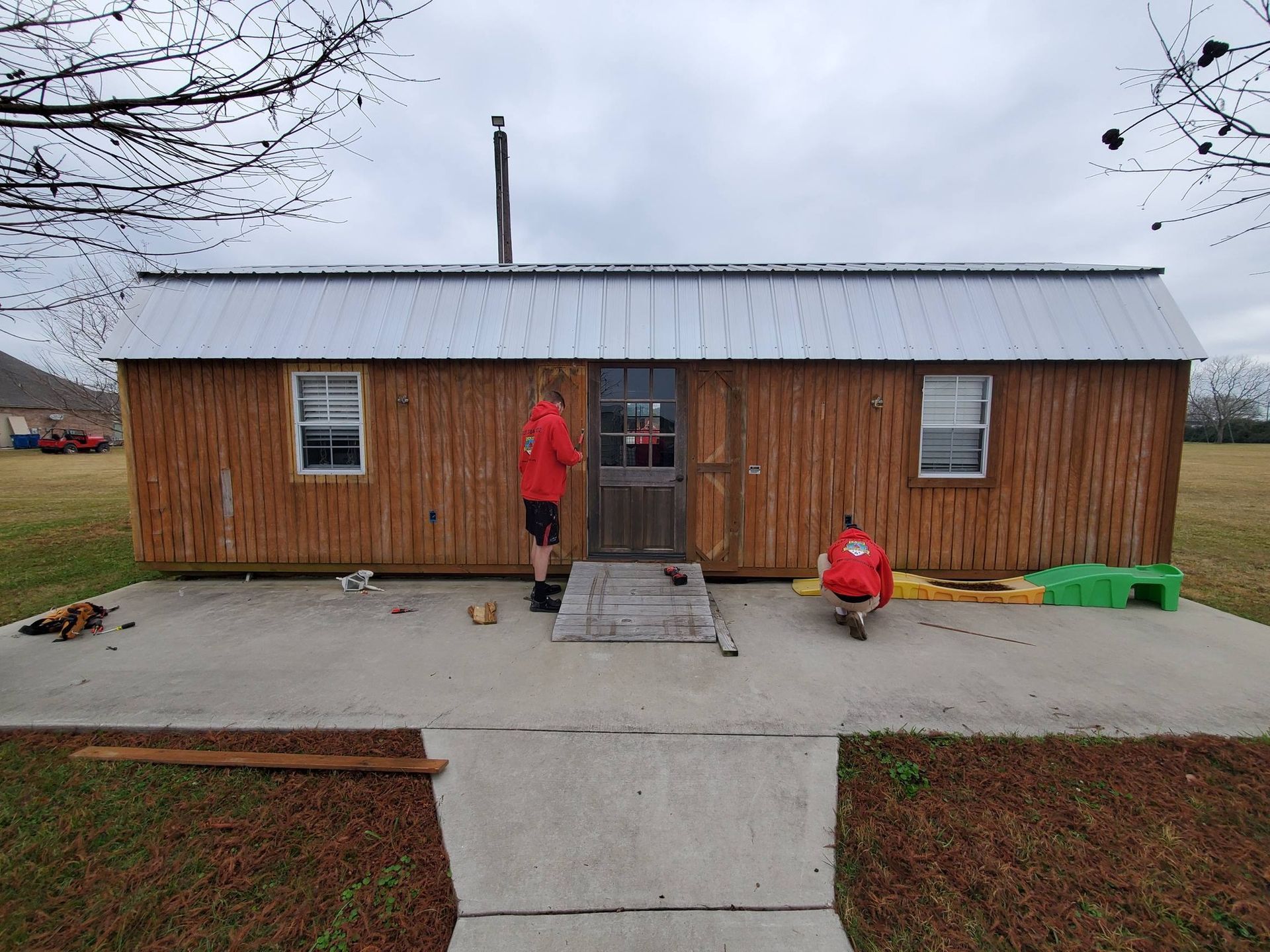 Two people in red jackets outside a small wooden cabin on a cloudy day