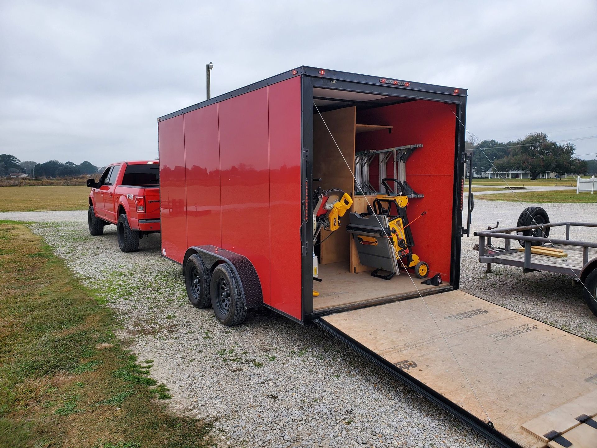 Red truck towing a red enclosed trailer with a yellow work machine inside on a gravel lot