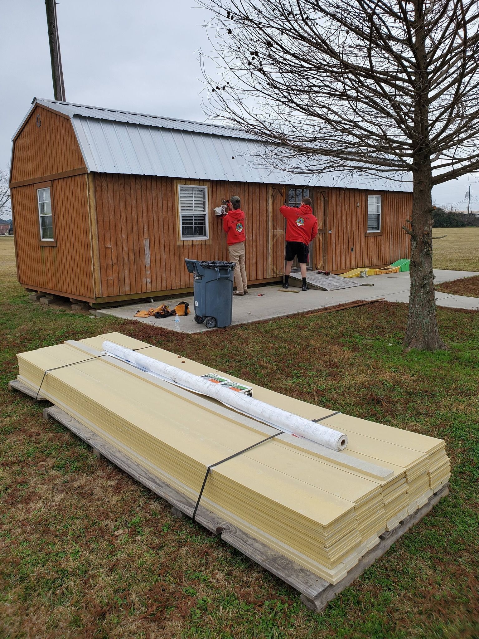 Two workers in red jackets stand beside a small wooden cabin on a rainy day.