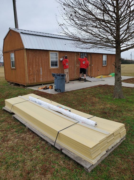 Two workers in red jackets stand beside a small wooden cabin on a rainy day.