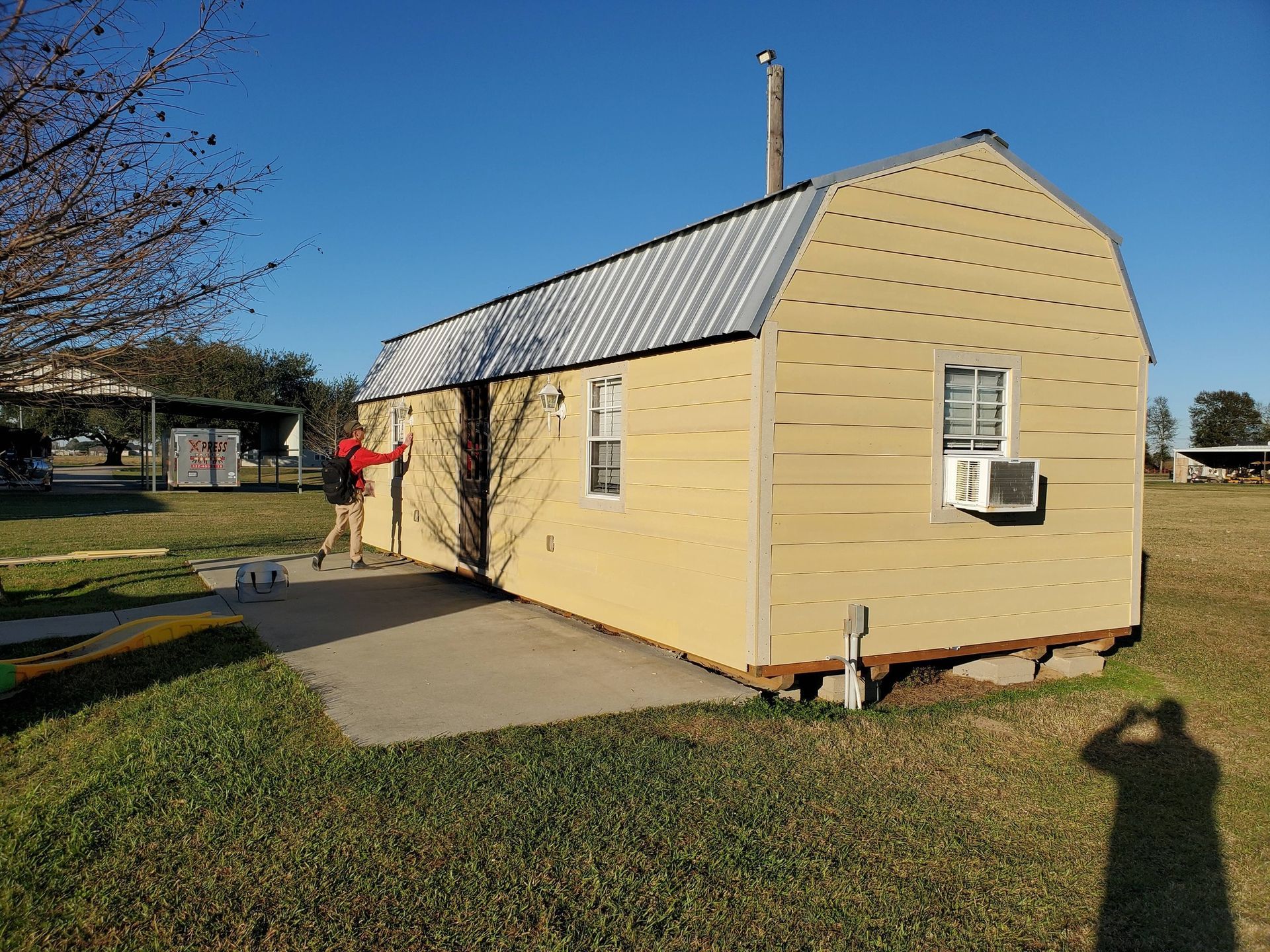 Small beige shed with a metal roof and window air conditioner beside a driveway in a grassy yard