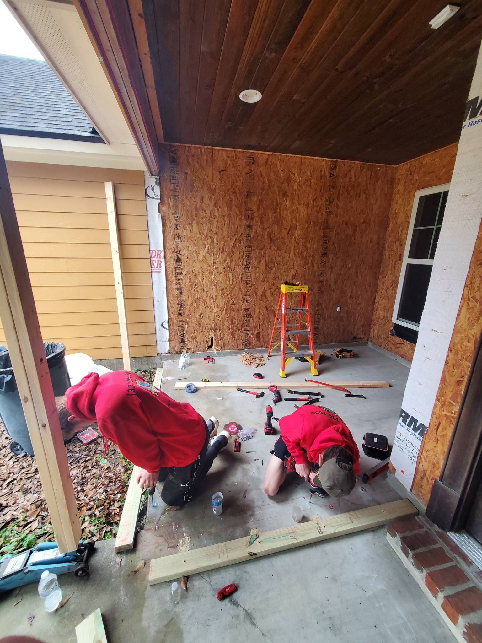 Two workers in red jackets crouch on a porch under construction, with tools and plywood walls.