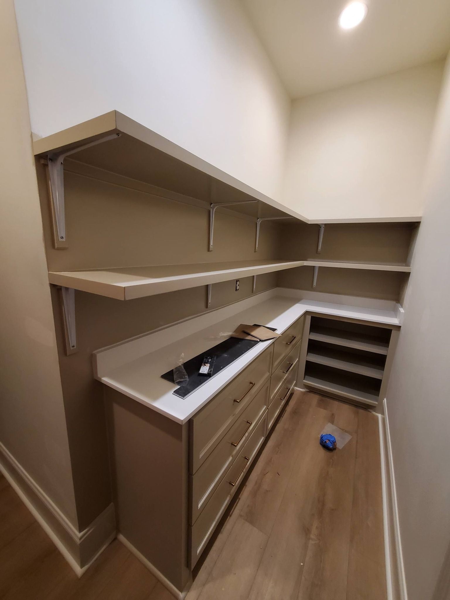 Empty walk-in pantry with brown shelves, wood floor, and a counter with tools and a blue bottle on the floor.