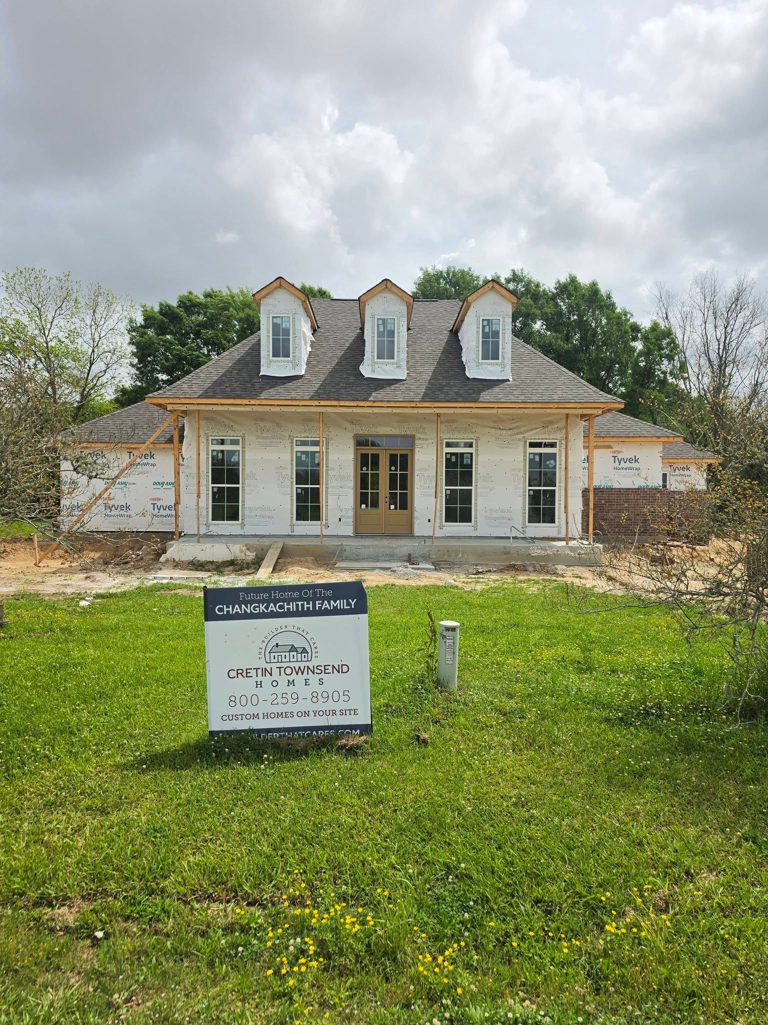 Small historic house with dormer windows behind a lawn and sign, under a cloudy sky.